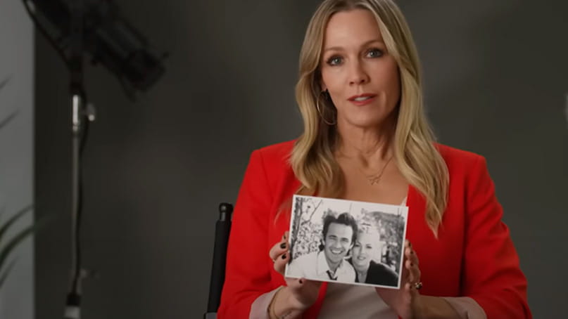 a video frame of Jennie Garth in an indoor setting holding a framed photo of herself with friend and fellow actor, Luke Perry