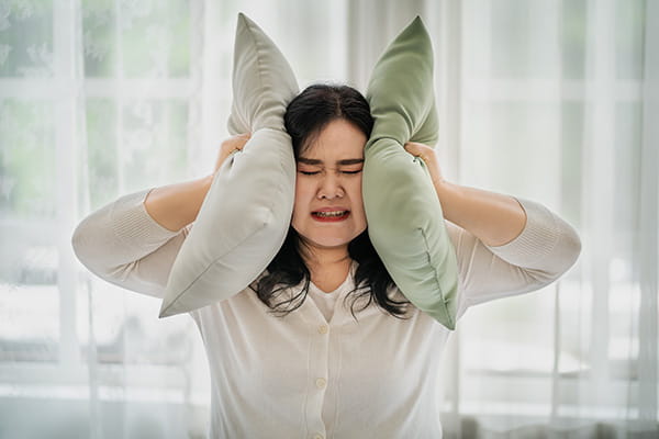 An Asian woman with a distressed expression is covering her ears with pillows.