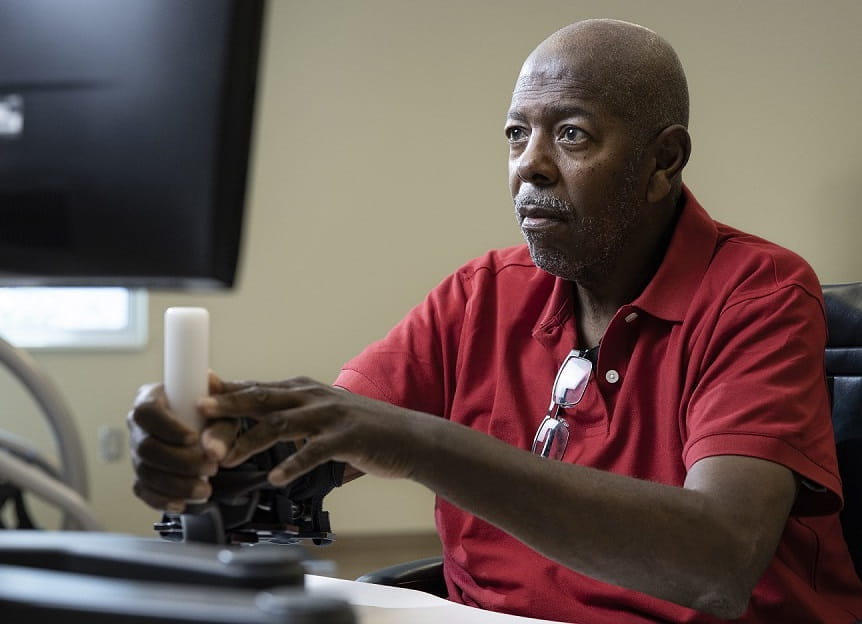 A senior Black man in a red polo is using a controller to interact with a computer.