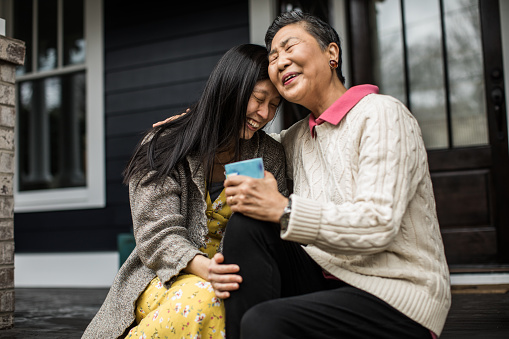 A senior Asian person and younger family member are sitting on a porch stoop embracing and smiling.