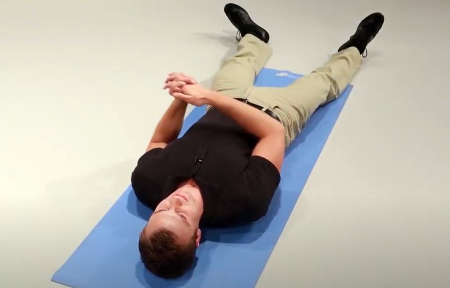 A White man is lying on his back on a mat with his legs outstretched and hands clasped.
