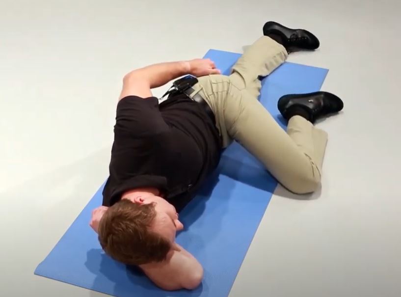 A White man is lying on his side with one arm supporting his head and his left knee bent and resting on the ground.