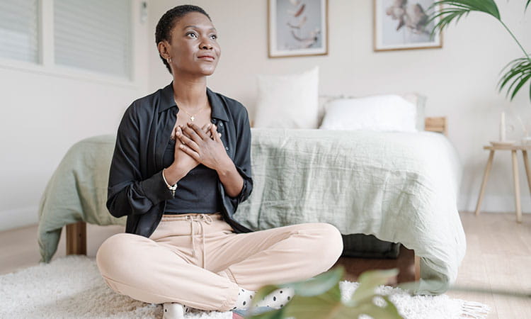 Woman with hands on her chest sitting on the floor in her bedroom in meditation.