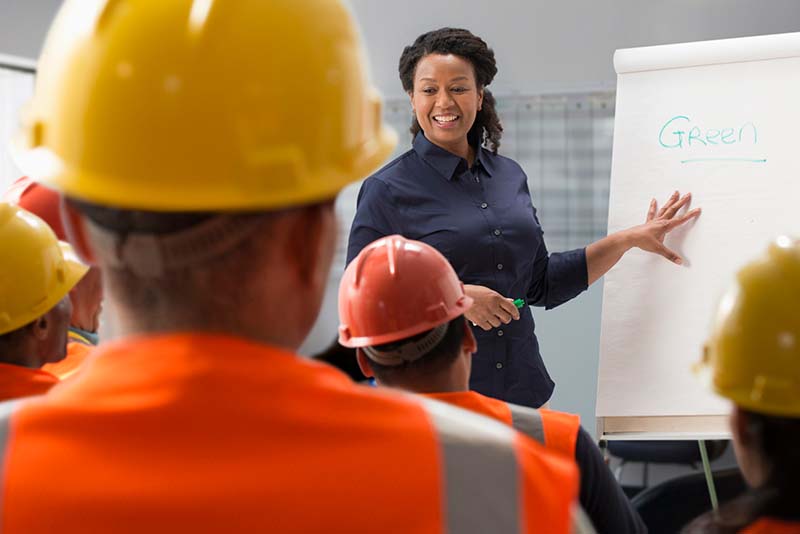 Woman pointing to white board, talking to employees wearing hard hats