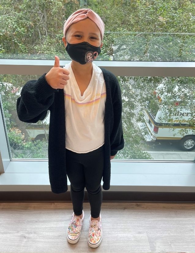 Pediatric stroke survivor Olivia Story wearing a mask standing and showing a thumbs up sign