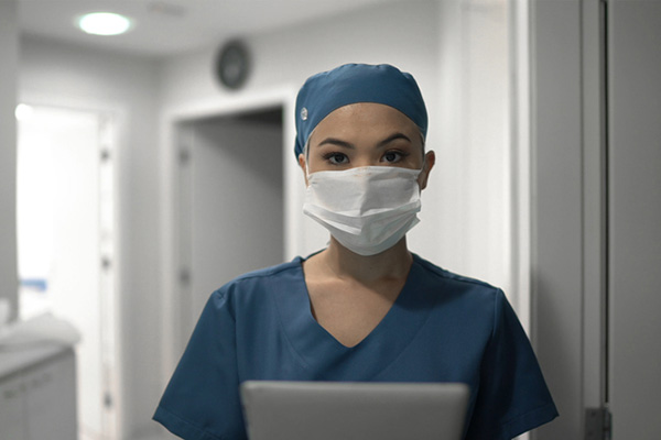 Masked female healthcare worker inside a hospital office area, holding a digital tablet