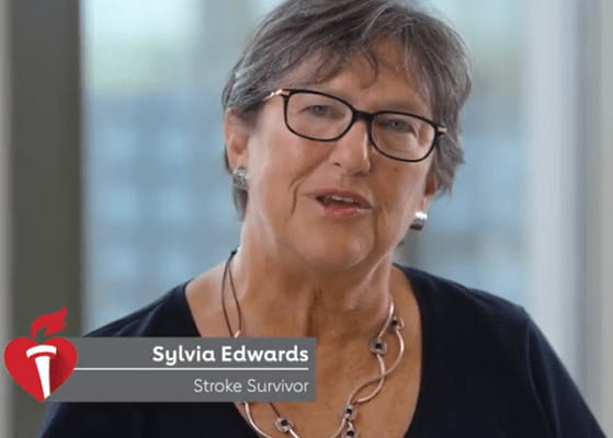 A video still of stroke survivor, Sylvia Edwards, speaking to the camera. She is a white woman with short, grey hair wearing black-framed glasses, a black top, a silver necklace and earrings.