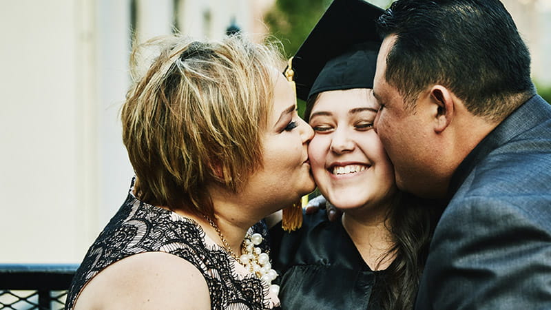 Hispanic/Latino mother and father kissing graduating daughter during family celebration