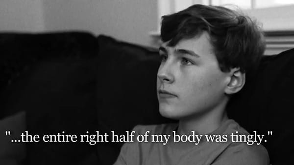A black and white video screen shot of stroke survivor, Alex, a white teenaged boy sitting on a sofa with the overlaying words, "...the entire right half of my body was tingly."