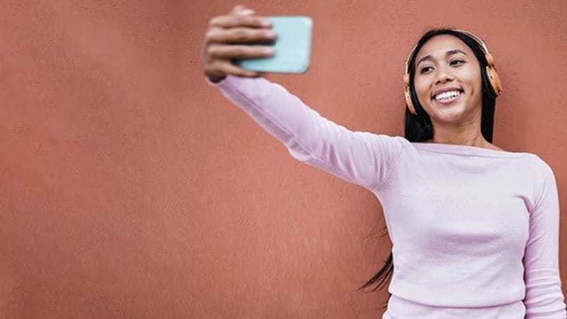 a young Black woman wearing headphones is smiling and taking a selfie with her smartphone against a terra cotta colored wall