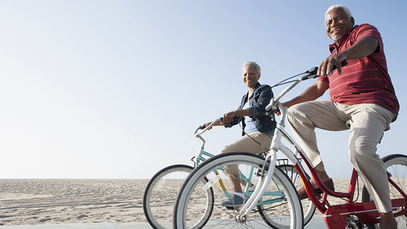 A senior Black couple is riding bicycles by a beach.