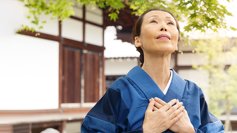 A mature Japanese woman wearing a blue kimono has her hands resting over her heart in a healing gesture as she looks up toward the sky.