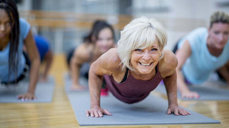 An older White woman is holding a plank pose and smiling while participating in a co-ed, multi-ethnic, fitness class.
