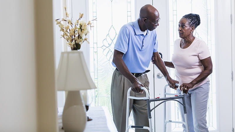 A senior Black couple inside their home at the front door. The man is using a mobility walker and his wife is helping him. They are in their 70s.