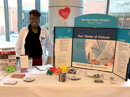 a woman representing Stamp Out Stroke is standing behind a table indoors with a Yale New Haven Hospital Stroke Center trifold display and other information materials