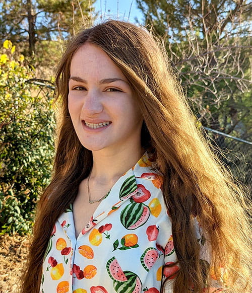 Mariah Fisher, a teenage girl with long hair, is smiling in an outdoor setting