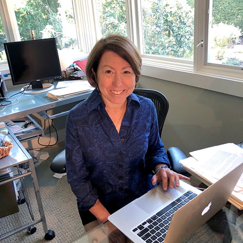 Debra Meyerson is looking up and smiling while sitting at a laptop in a home office setting