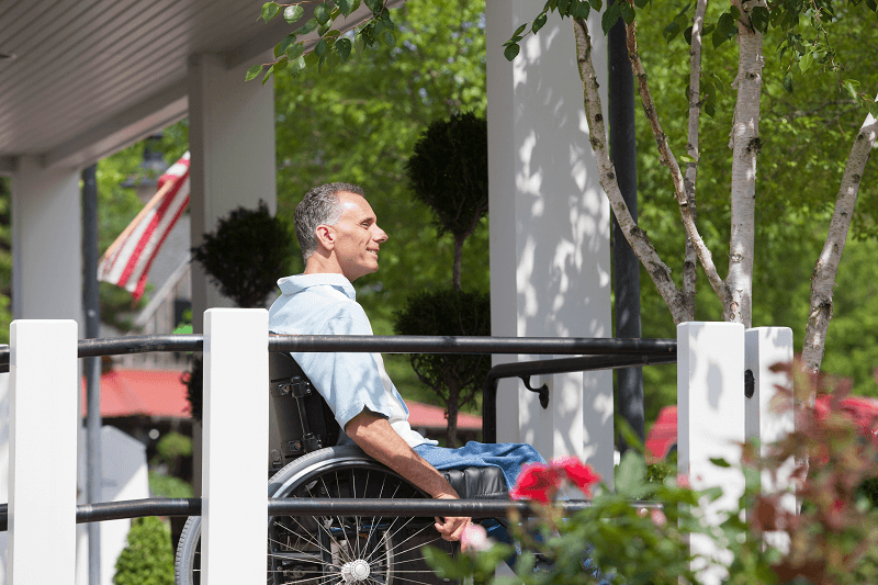 A mature man is sitting in a wheelchair outside on a home porch surrounded by trees.