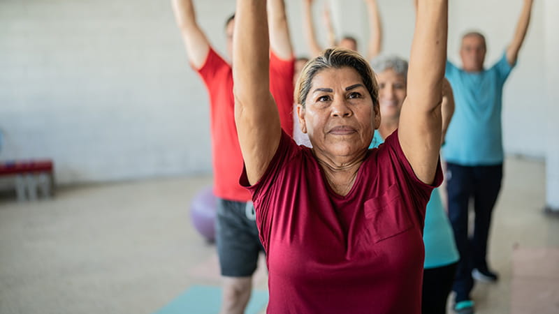 A senior woman is stretching upward with classmates in a yoga studio.