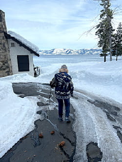 Deb Meyerson is wearing a coat and hat and walking with a walking stick on a snowy drive with Lake Tahoe and mountains in the background