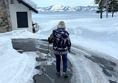 Deb Meyerson is wearing a coat and hat and walking with a walking stick on a snowy drive with Lake Tahoe and mountains in the background