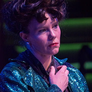 Maggie Whittum is wearing a teal sequined jacket and has an expression of sadness on her face while acting out a scene in a play.