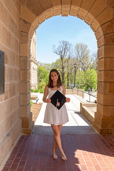 Hannah Halaska holding a graduation cap under a building archway (© Phyllis Bankier)