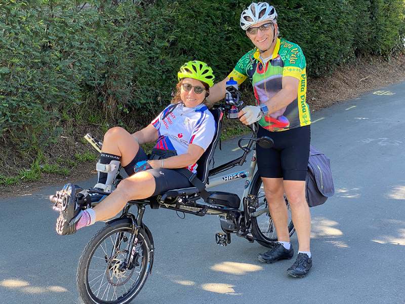Debra Meyerson and Steve Zuckerman are dressed in cycling gear and posing with their tandem bike in an outdoor setting.