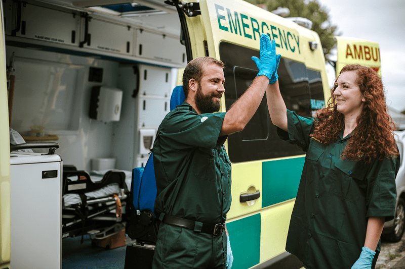 Two smiling paramedics are high-fiving outside of an emergency vehicle.
