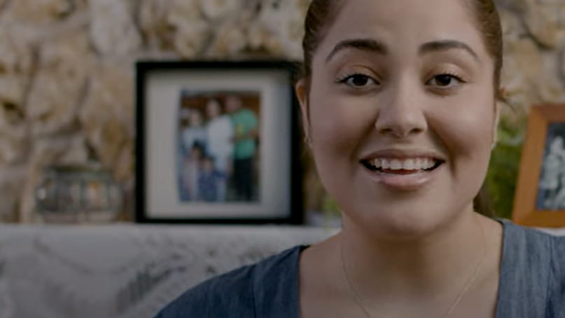 A young Hispanic/Latina woman is smiling in front of a framed family photo sitting on a fireplace mantel.