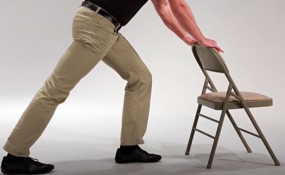 A White man is standing in a lunge position with this hands resting on the back of a chair for support.