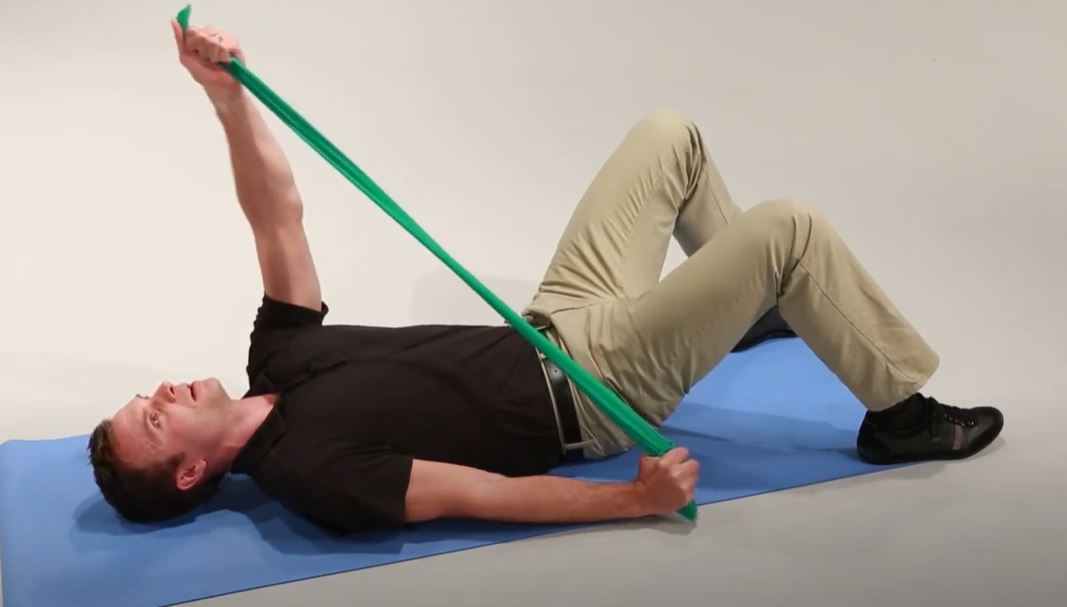 A White man is lying on his back on a mat with his knees bent and feet flat on the ground. His arms are extended as he pulls each end of a resistance band.