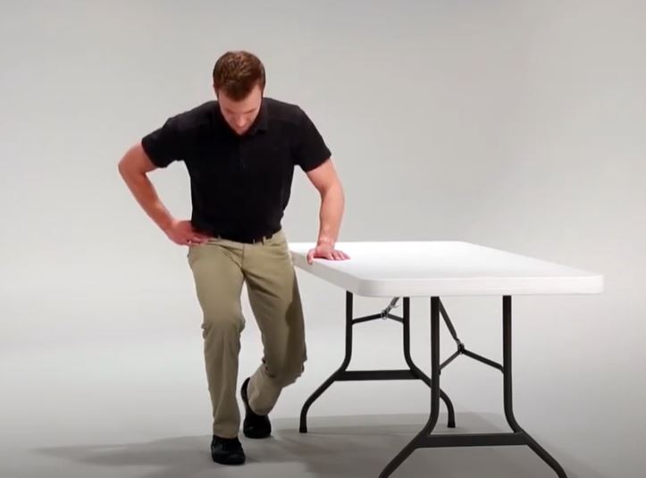A White man is kneeling with his left hand on a folding table for support.