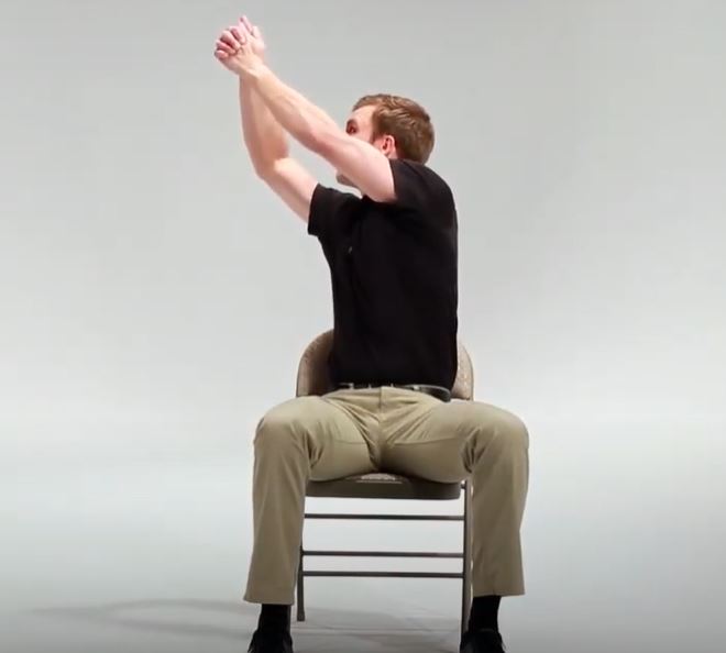 A White man is sitting in a chair reaching is arms up and over to his right side.