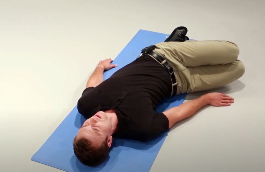 A White man is lying flat on his back on a mat with his knees bent and rotated to the right.