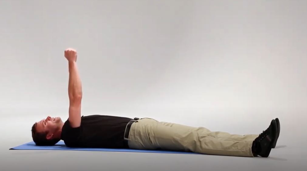 A White man is lying flat on his back on a mat with his arms extended straight up in the air.