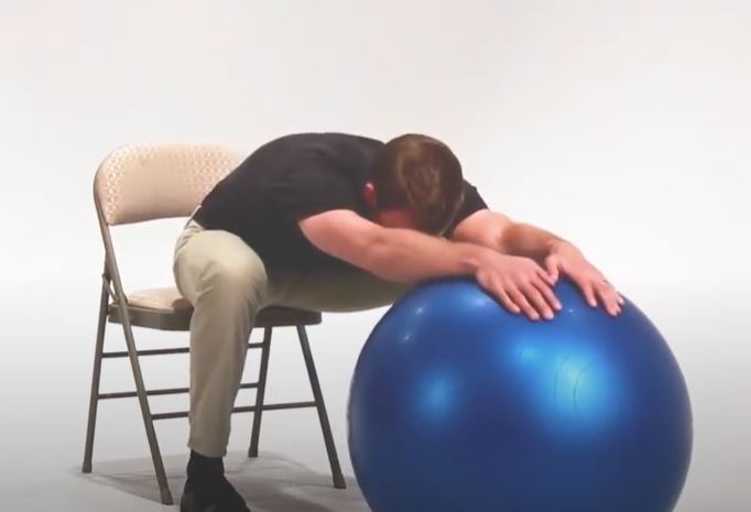 A White man is sitting in a chair and stretching forward over a large exercise ball.