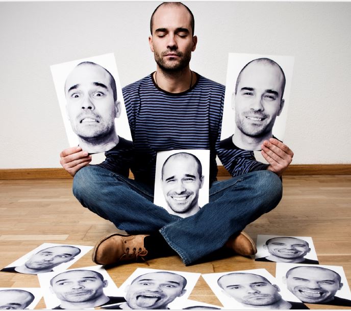 A man is sitting cross-legged on the ground surrounded by scattered black and white print-outs of his face with various expressions.
