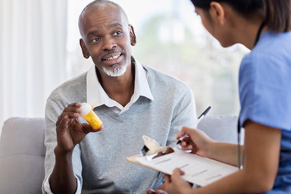 a middle-aged Black man showing a bottle of prescription medication to a female medical professional taking notes on a clipboard