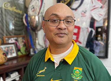 Stroke survivor, Silivenusi “Nusi” Tukuafu, is standing in front of sports memorabilia wearing a Springboks rugby jersey.