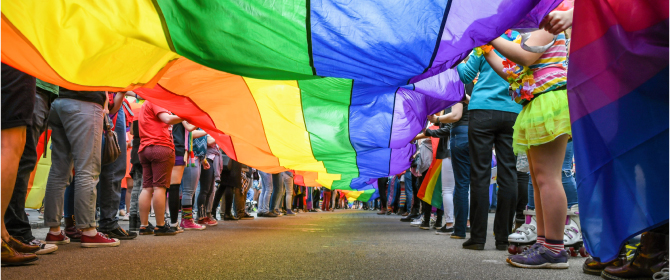 A view from under a long rainbow fabric held up by two lines of people on a city street. A view from under a long rainbow fabric held up by two lines of people on a city street.