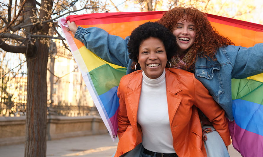 A person is giving a piggyback ride to another person holding a rainbow flag in an urban park setting. A person is giving a piggyback ride to another person holding a rainbow flag in an urban park setting.