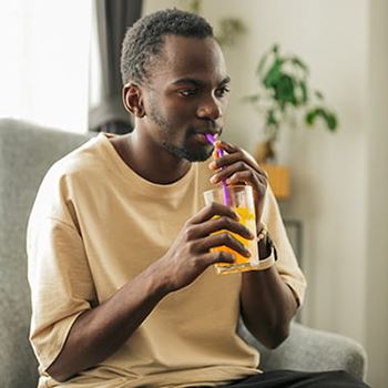 Black man drinking iced drink through a straw sitting on the sofa at home