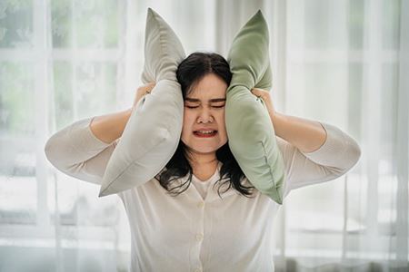 An Asian woman with a distressed expression is covering her ears with pillows. An Asian woman with a distressed expression is covering her ears with pillows.