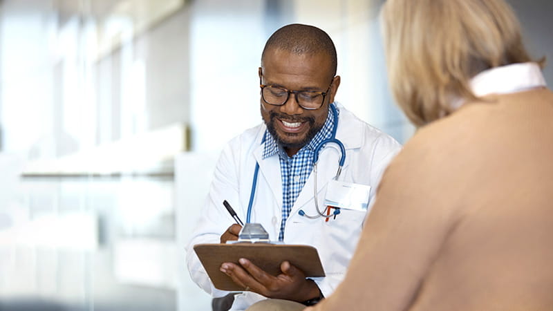 A smiling Black doctor is taking notes while talking with his senior female patient. A smiling Black doctor is taking notes while talking with his senior female patient.