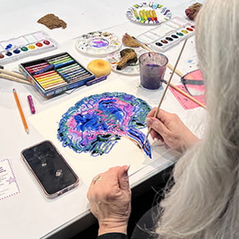 An over-the-shoulder view of an older woman painting an abstract brain on a canvas An over-the-shoulder view of an older woman painting an abstract brain on a canvas
