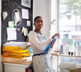 A Black woman is standing in an office next to a large window holding a clipboard. woman in art studio holding clipboard