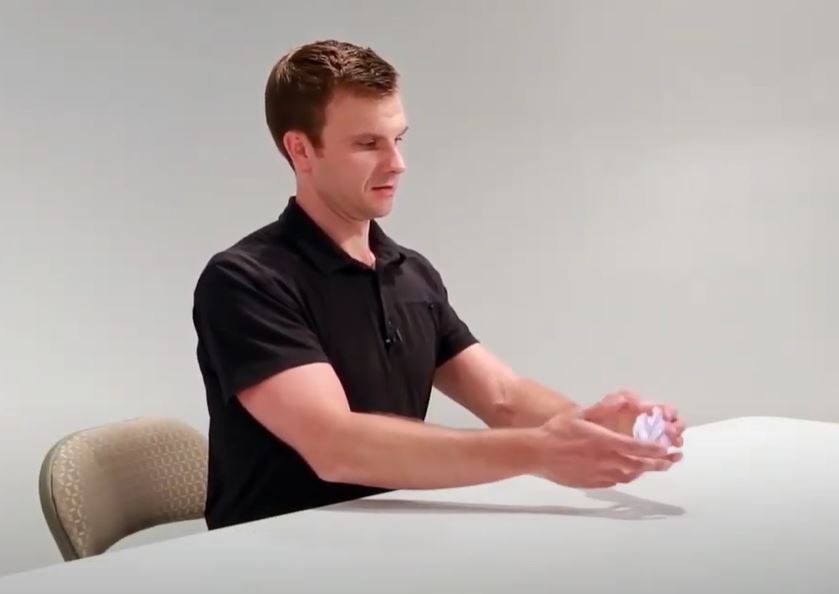 A White man is sitting at a table with his arms outstretched in front of him. His hands are holding a crumpled ball of paper.