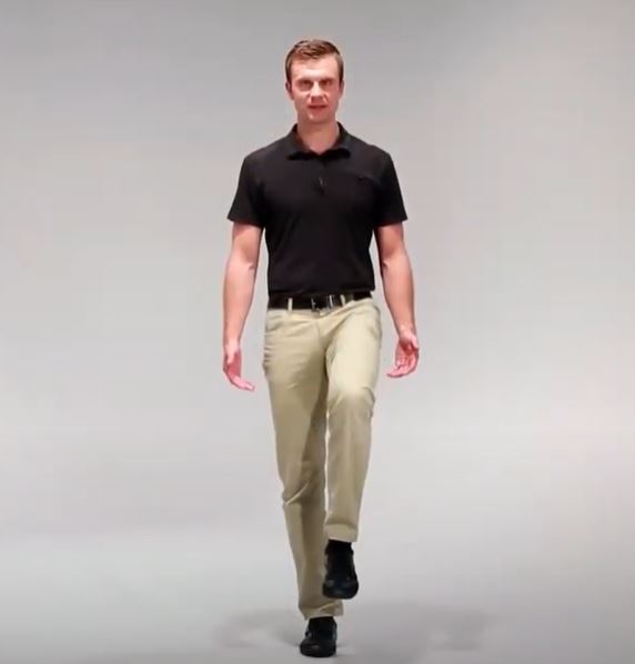 A White man is standing straight with his left knee bent and lifting his leg off the ground.