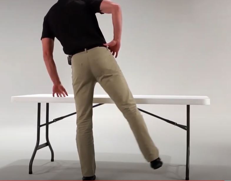 A White man is leaning on his left leg with his right leg out to his side. His left hand is resting on a folding table for support.